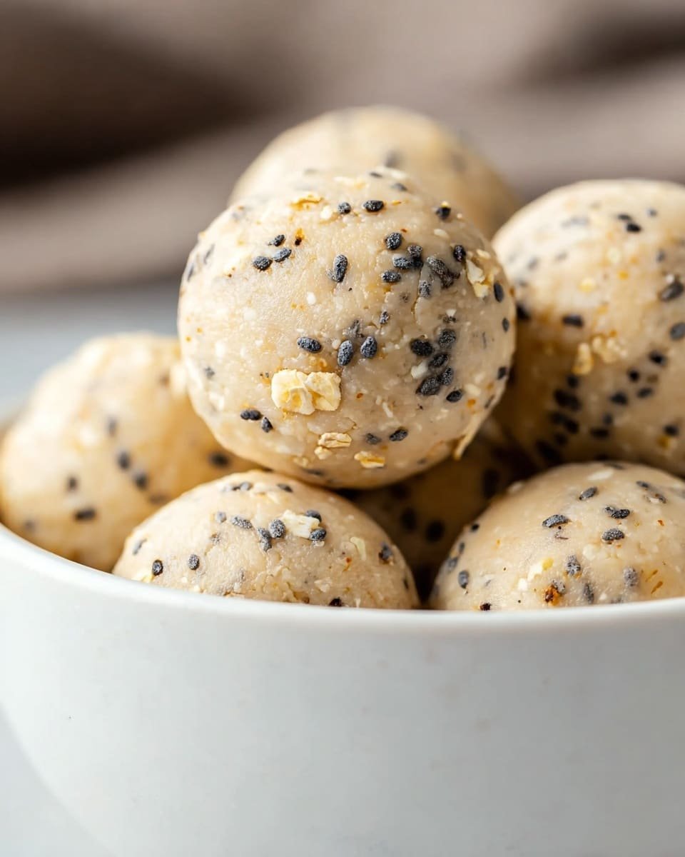 A close-up of several round energy balls inside a white bowl, each ball showing a pale beige base speckled with small black seeds and visible light oat flakes, giving the surface a slightly rough texture with tiny bumps; the balls are stacked loosely, filling the bowl almost to the top, with a smooth white marbled background softly blurred in the back, the lighting soft and natural highlighting the texture of the balls and oats, photo taken with an iphone --ar 4:5 --v 7