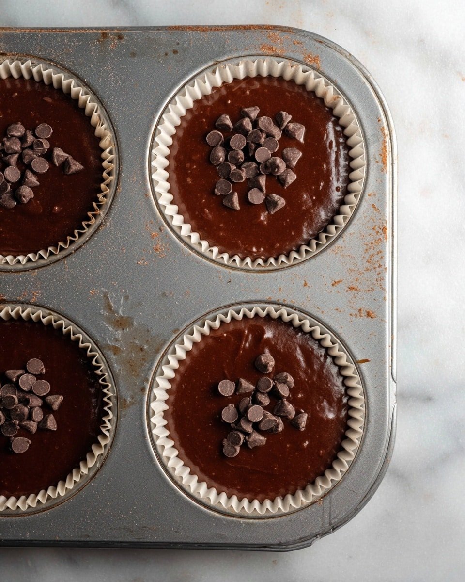 A metal muffin tray holds three white paper cupcake liners filled with smooth, dark chocolate batter. Each chocolate layer is glossy and rich, topped with a small cluster of dark chocolate pieces scattered unevenly. The tray shows light signs of use, with tiny stains and marks. The background is a white marbled texture, making the chocolate batter and cupcakes stand out clearly. photo taken with an iphone --ar 4:5 --v 7