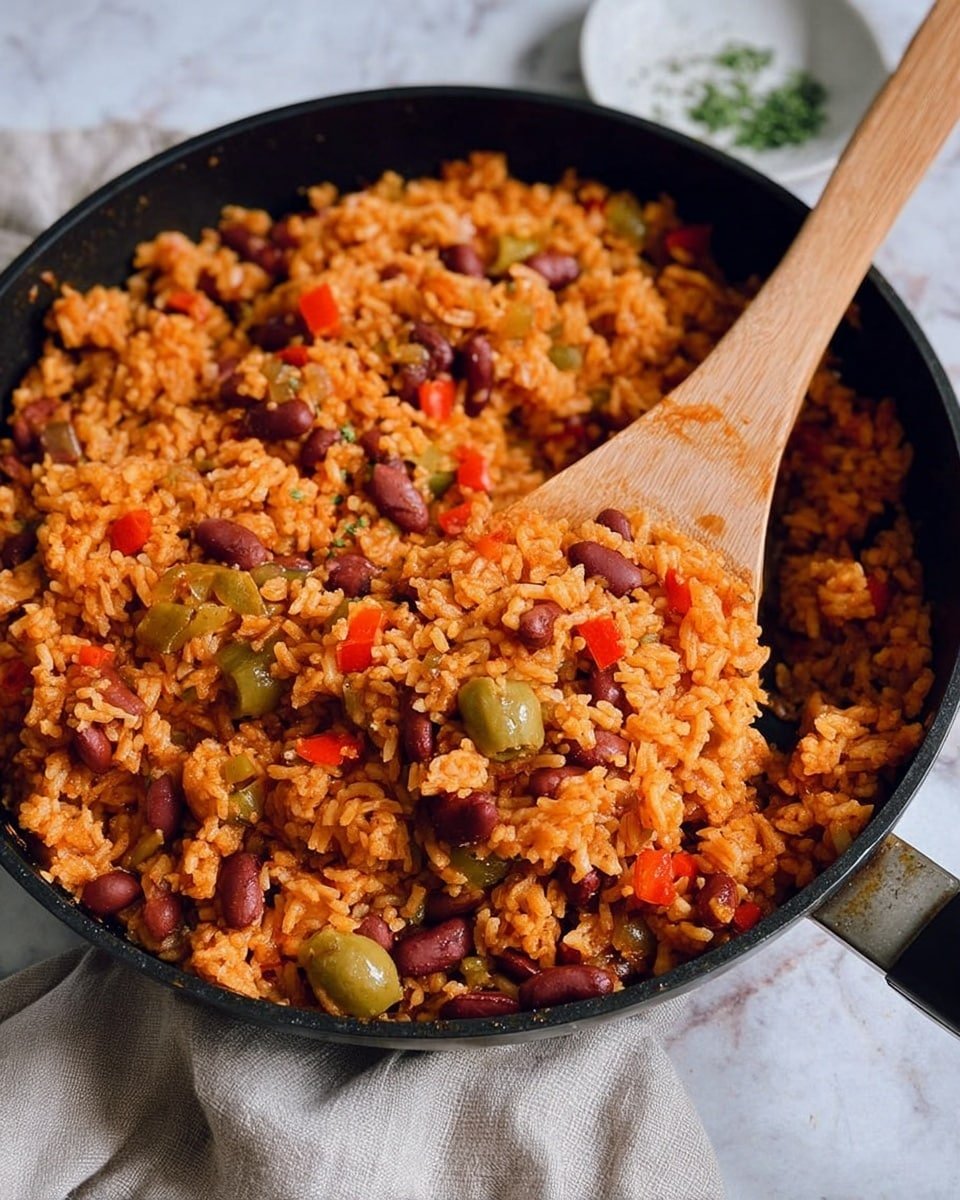 A close-up of a black pan filled with a mix of cooked orange rice, red beans, diced red bell peppers, green olives, and small bits of onions, all mixed together with a moist texture. A wooden spoon is resting inside the pan, scooping some of the rice mixture. The pan is placed on a white marbled surface with a light grey cloth and a small white bowl partially visible around it. photo taken with an iphone --ar 4:5 --v 7